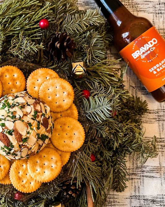 Cheese ball coated in almonds with crackers, pine wreath décor, and Bravado Spice hot sauce in a Christmas-themed setting.