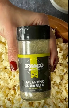 A hand sprinkling Bravado Spice Co.’s Jalapeño & Garlic Seasoning over a bowl of freshly popped popcorn.