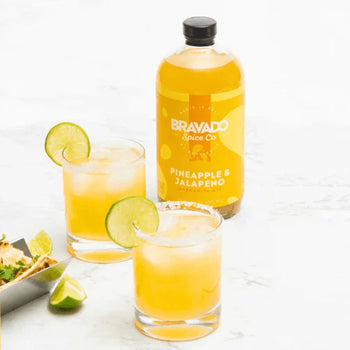 Bravado Spice Co.’s Pineapple & Jalapeño margarita mixer with two cocktails in glasses and fresh lime slices on the side.