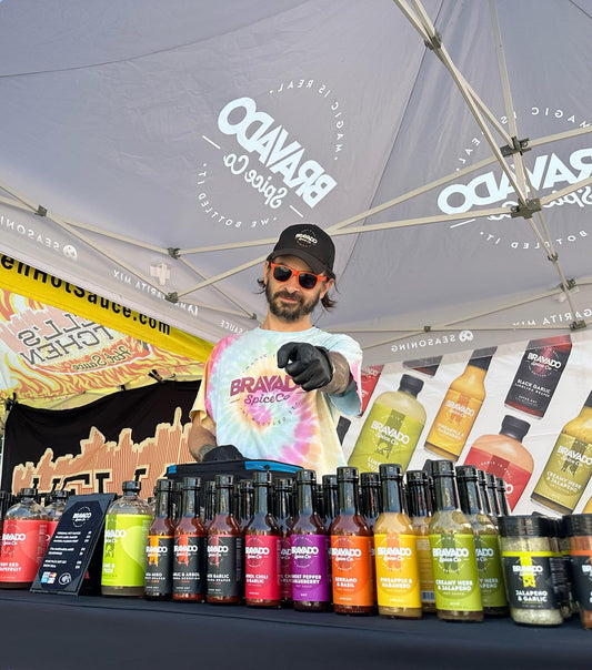 A man at a Bravado Spice Co booth points at the camera behind rows of hot sauce bottles.