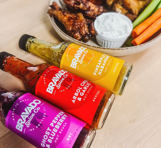 Three bottles of Bravado Spice hot sauce in front of a plate of chicken wings, dip, celery, and carrot sticks.