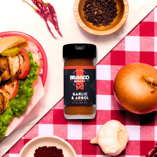 A jar of Bravado Spice Co. Garlic & Árbol seasoning surrounded by ingredients and a sandwich.