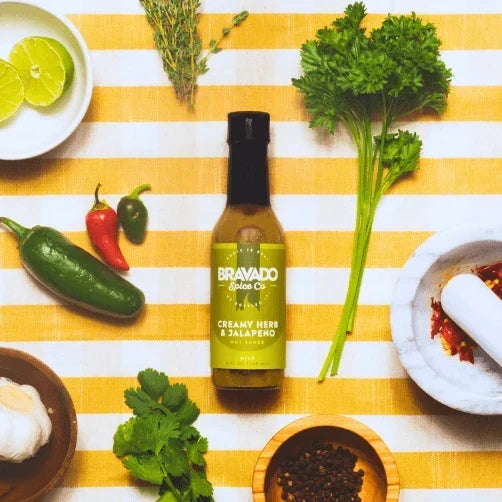 Bravado Spice Creamy Herb & Jalapeno Hot Sauce surrounded by fresh herbs, peppers, and limes.