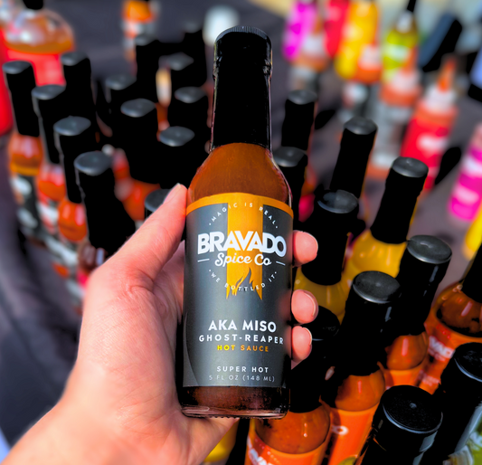  A hand holding AKA Miso Ghost-Reaper hot sauce over many other hot sauce bottles.