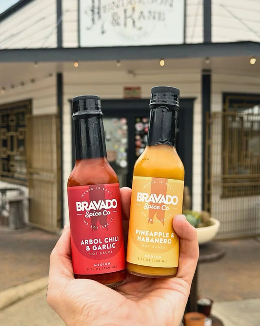 A hand holds Bravado Spice Arbol Chili & Garlic and Pineapple & Habanero hot sauce bottles outside a Texas store.