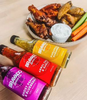 Three Bravado Spice sauces alongside a plate of chicken wings, dip, and fresh celery sticks.