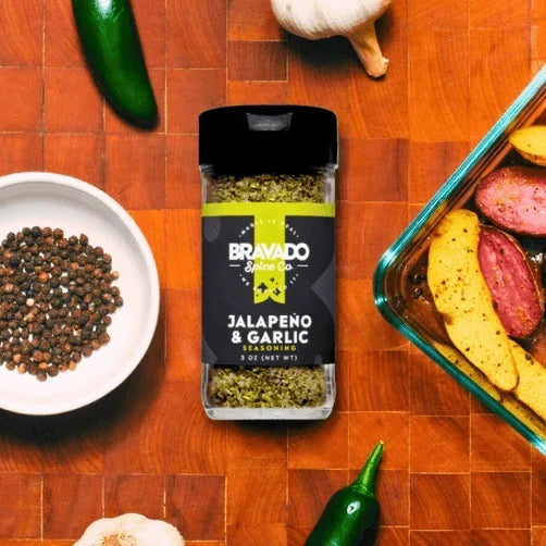 Bravado Spice Jalapeño & Garlic Seasoning bottle next to potatoes and fresh ingredients for a cooking scene.