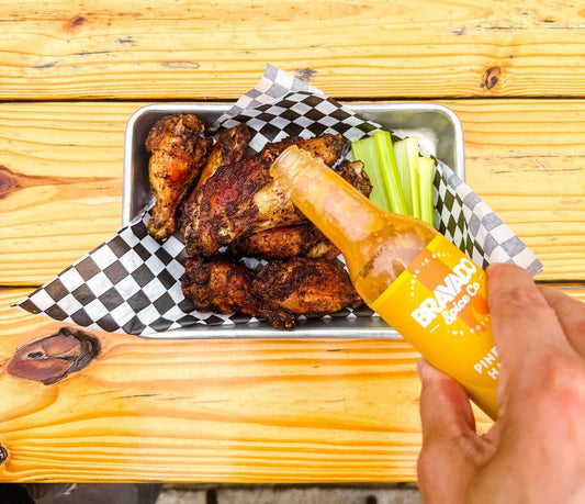 A hand uses Bravado Spice Co.’s Pineapple and Habanero hot sauce on a tray of wings.