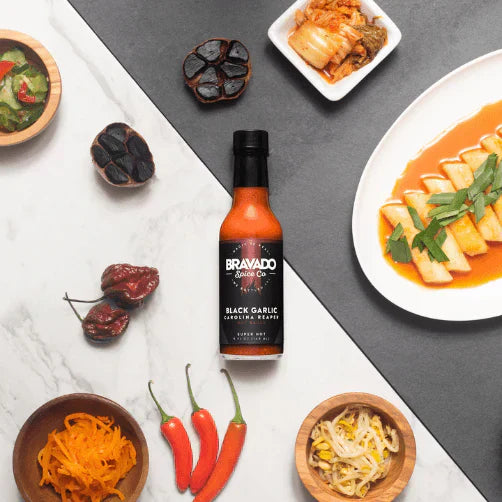 Bottle of Bravado Spice Company Black Garlic & Carolina Reaper Hot Sauce surrounded by spices, peppers, and plated dishes.