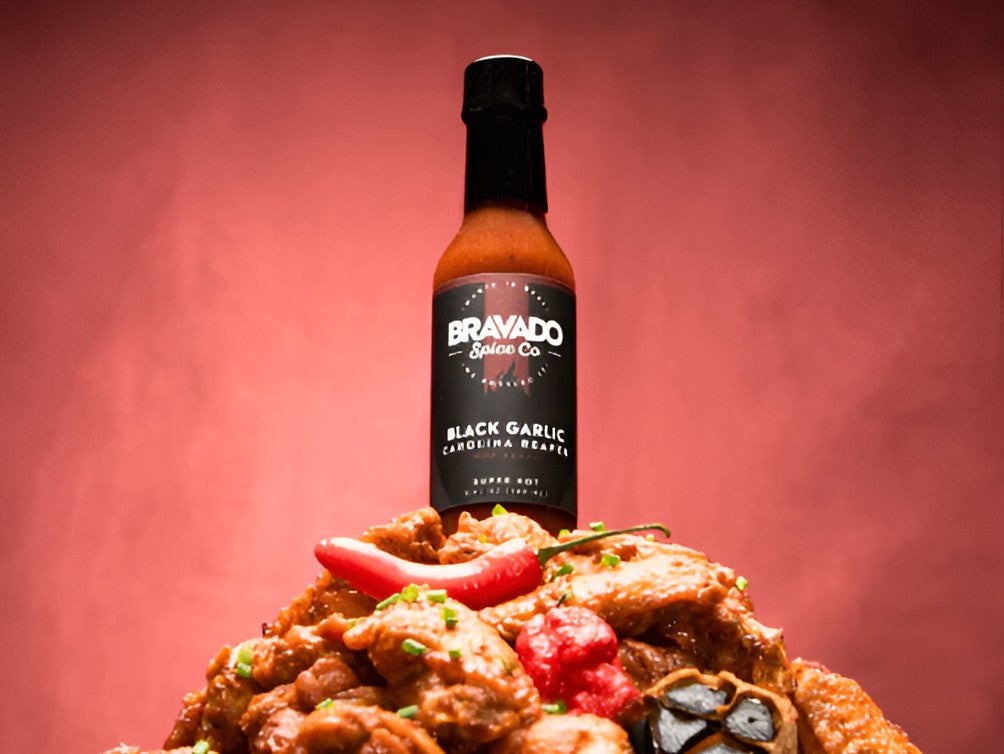 Bravado Spice Co Black Garlic Carolina Reaper hot sauce bottle set atop a savory plated dish with red chili peppers and herbs against a bold red background.